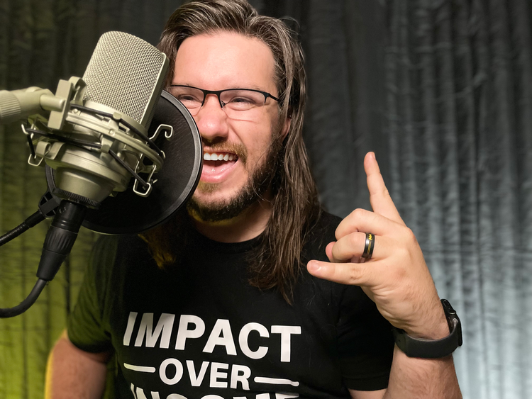 A young white man with long brown hair and a trimmed beard and mustache stands behind a boom mic. He is wearing glasses and a black t-shirt. He is grinning at the camera and throwing up the horns.